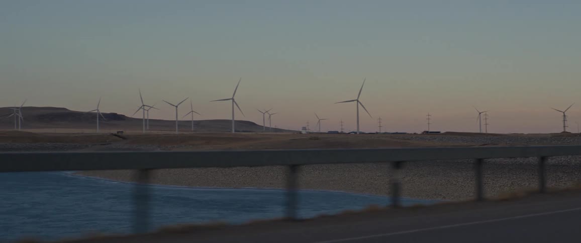 Bridge crosses over blue water contrasting with brown and yellow grass on hill leading to spinning turbines on wind farm against the red tinted sky off the horizon