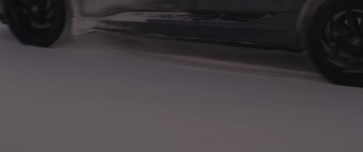 A black car travels on a snow covered road and snow is sticking to the car