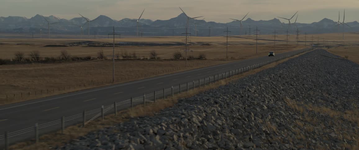 A vehicle driving on a road with wind turbines a mountain range the sky and clouds in the background