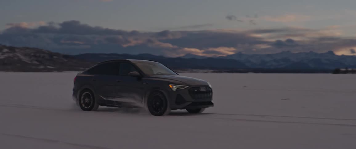 A black car travels through the snow with snow covered mountains in the distance