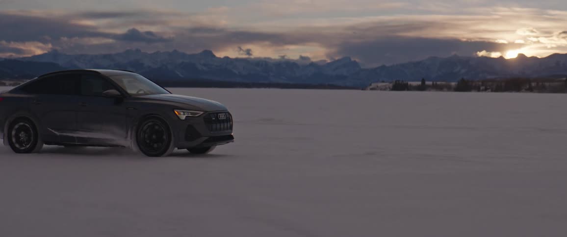 A black car travels through the snow as the sun is setting with mountains in the distance