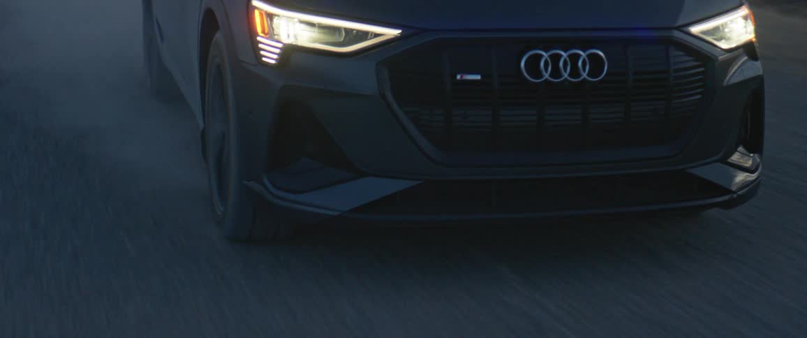 Front end view of a black audi automobile traveling on a road and leaving a trail of smoke
