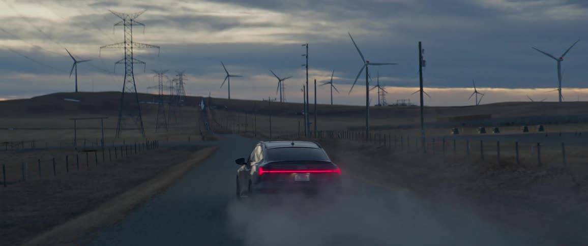 A car travels a road beside a group of wind turbines kicking up dust