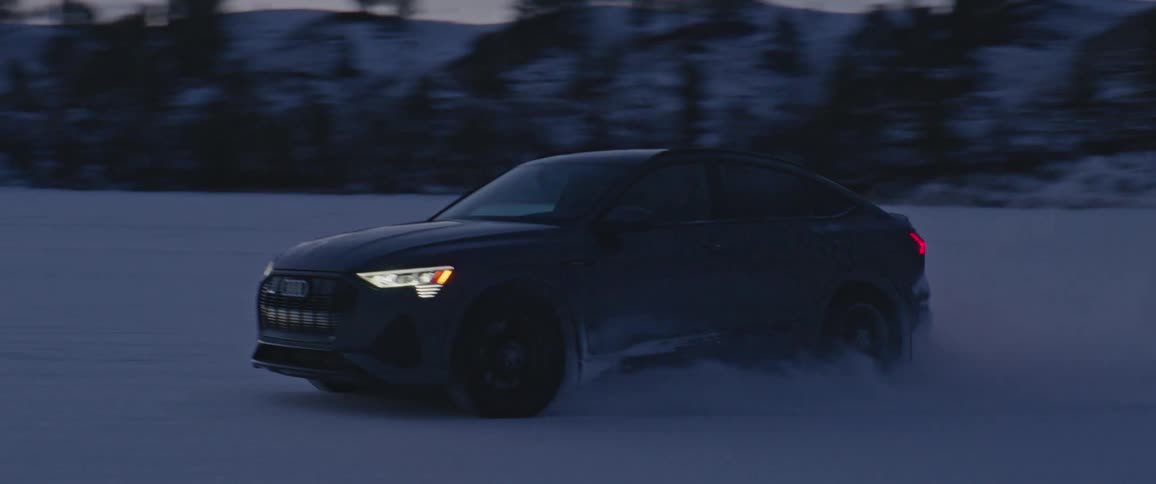A black car with headlights on travels on a snow covered road with mountains and trees in the distance