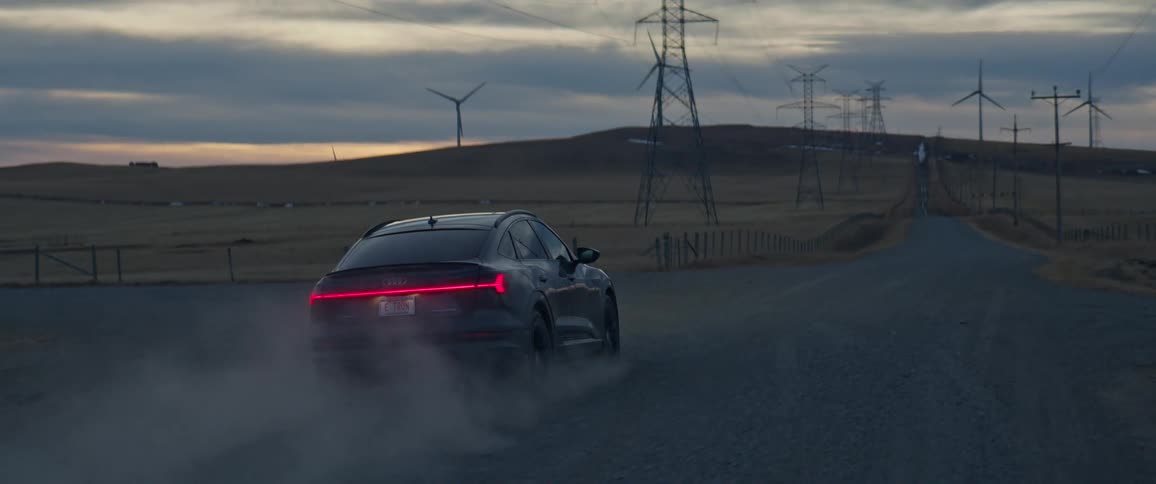 A black car travels on a road with spinning wind turbines and utility towers along the roadside