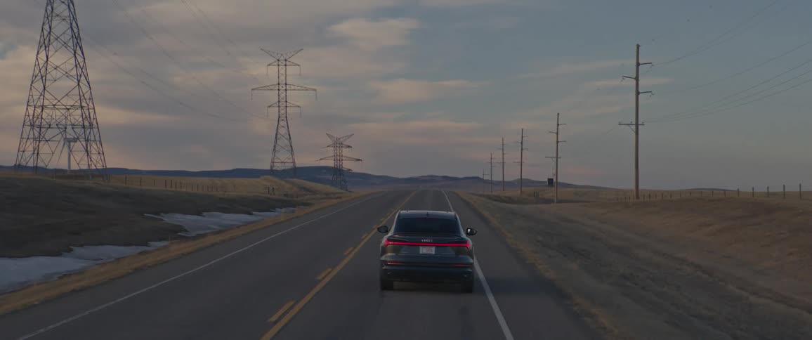 A black car travels on a road with wind turbines and utility towers along the roadside