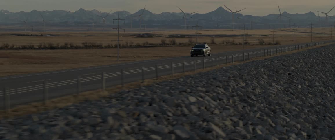 A black vehicle drives down a rural highway near a canyon and windmills