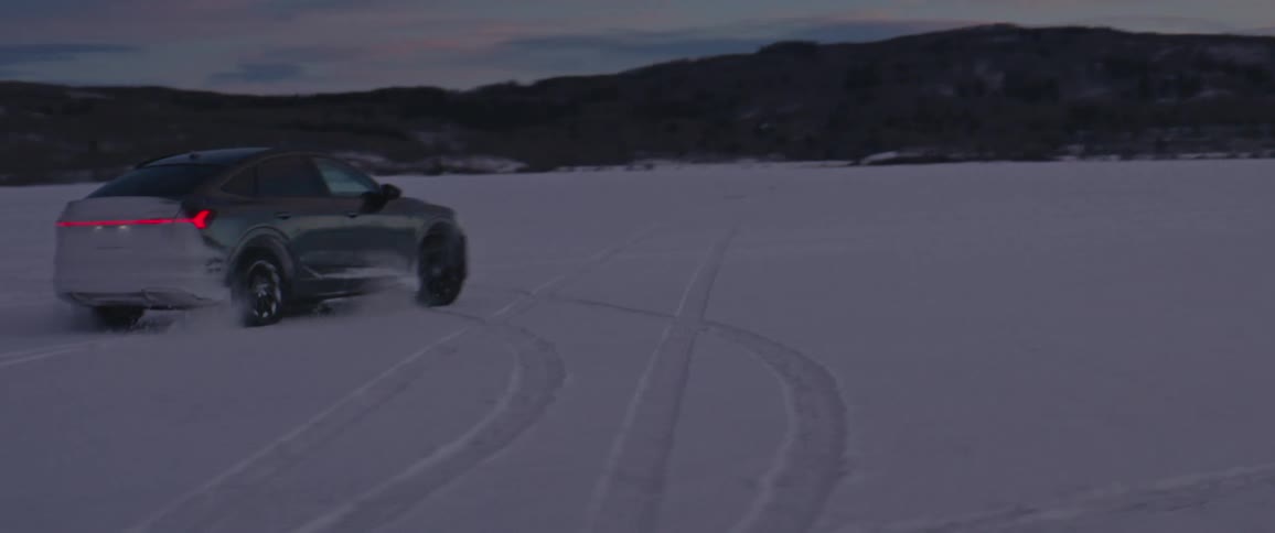 A black car travels in circles on a snow covered road with mountains in the distance
