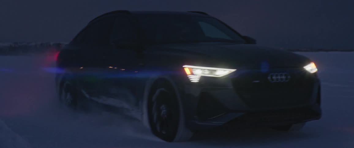 Car speeds across snowy plain away from buildings at night kicking up snow
