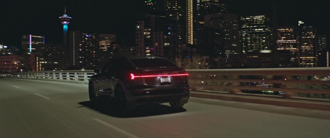 A black audi car is driving on a flyover with tall buildings in sight