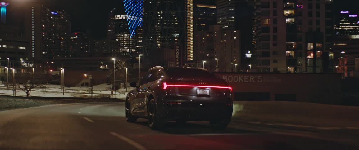 Dark grey audi car drives on city highway with illuminated high rise buildings in the background