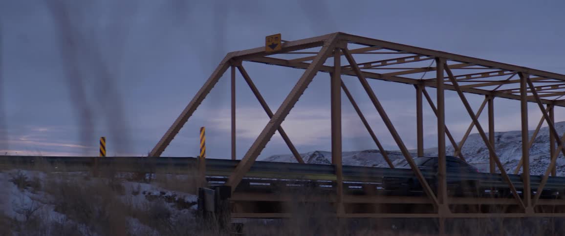 A car drives across a bridge in front of snowy hills