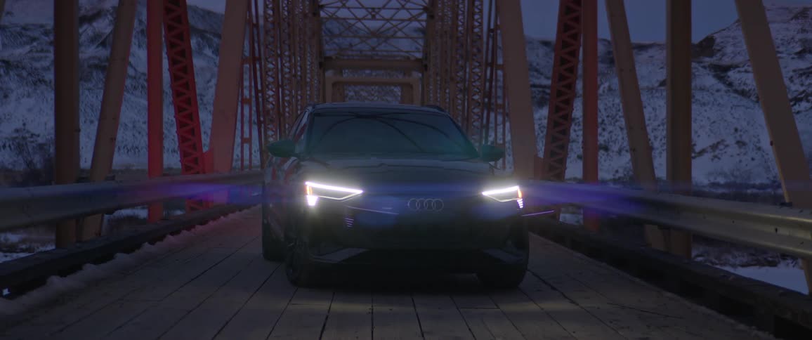 Black audi with its headlights on sits parked on wood and metal bridge over icy river with snow covered hills and mountains in background