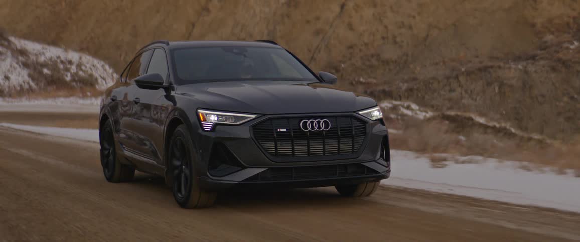 A black audi automobile travels on a road with snow along the roadside and mountains in the distance