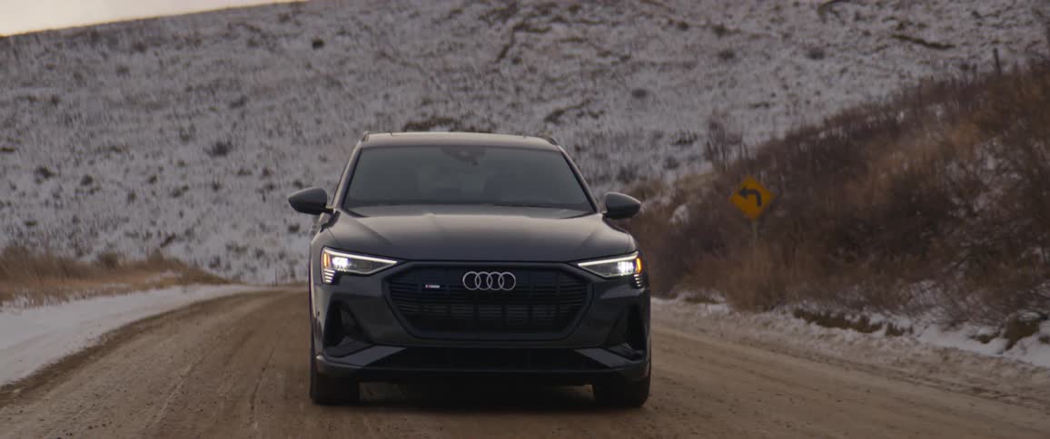 A black audi automobile travels on a road with snow covered mountains in the distance