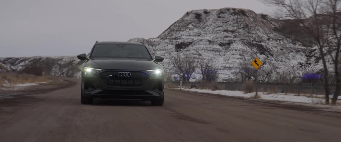 A black car with headlights on travels on a road with snow covered mountains in the distance