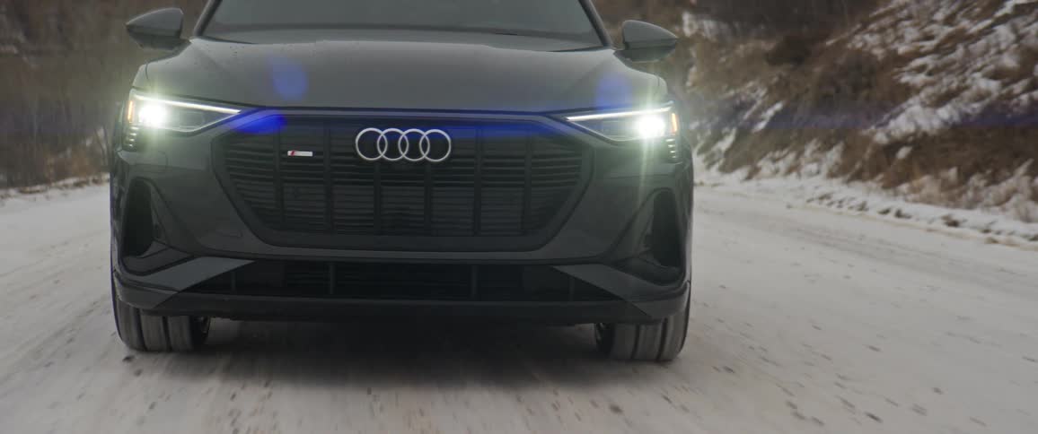 A black audi car with headlights on travels on a snow covered road with mountains in the distance