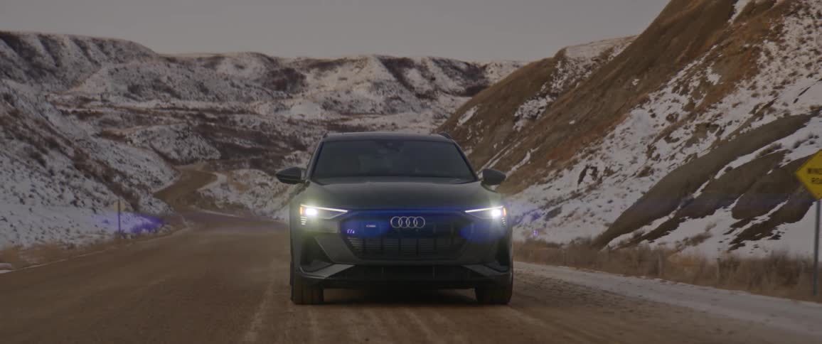 A vehicle travels a dirt road beside snow dusted mountains