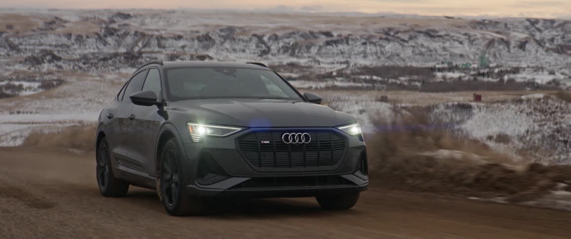 A black audi automobile travels on a road with snow covered mountains in the distance