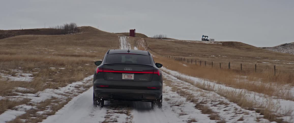 Car drives along snow covered dirt road past fenced pastures toward oil pump sitting on top of hill