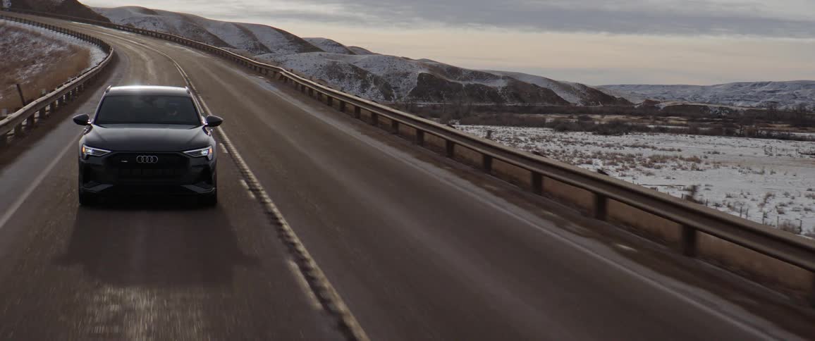 Dark grey audi car drives downhill on highway with snowy hills in the background