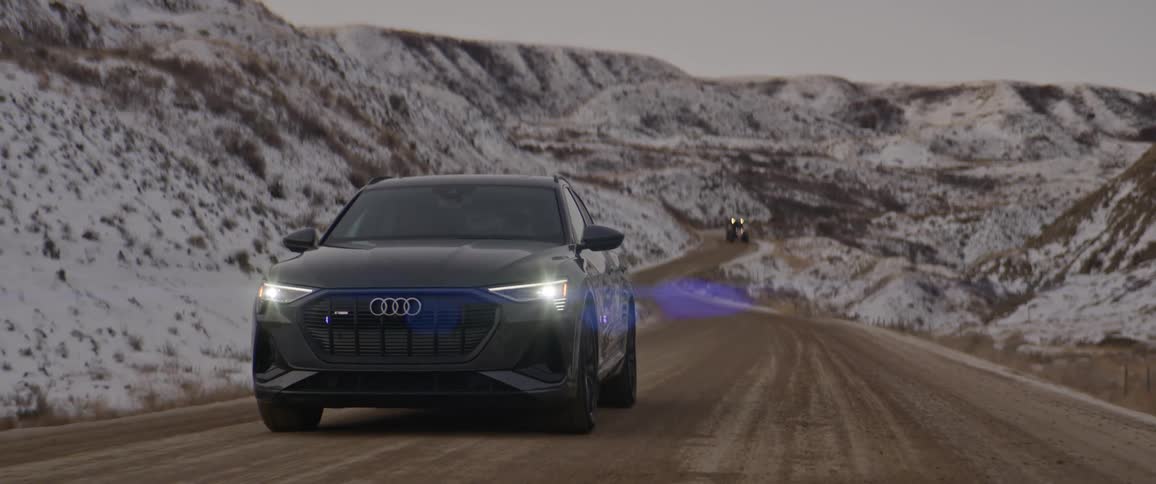 A dark sedan drives up a wet mountain road that cuts through snowy hills