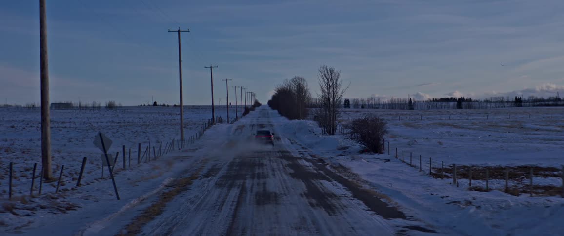A black car travels on a snow covered road with trees and utility poles along the roadside