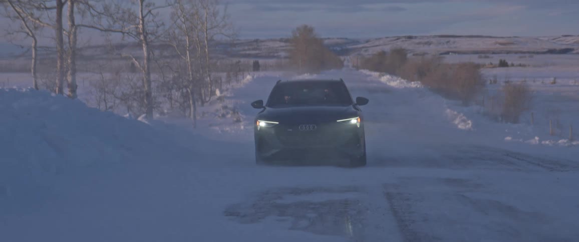 A black audi automobile travels on a snow covered road with trees along the roadside