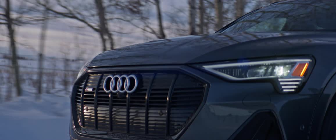 The front end of a black audi traveling on a snowy road with trees in the distance