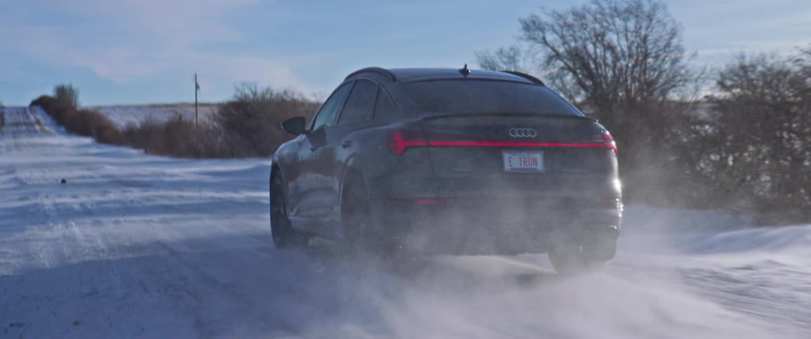 A black car travels on a snow covered road with trees along the roadside