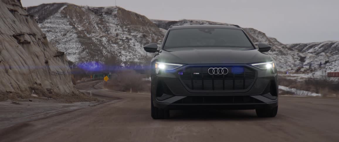 Dark audi car drives uphill with snowy hills in the background