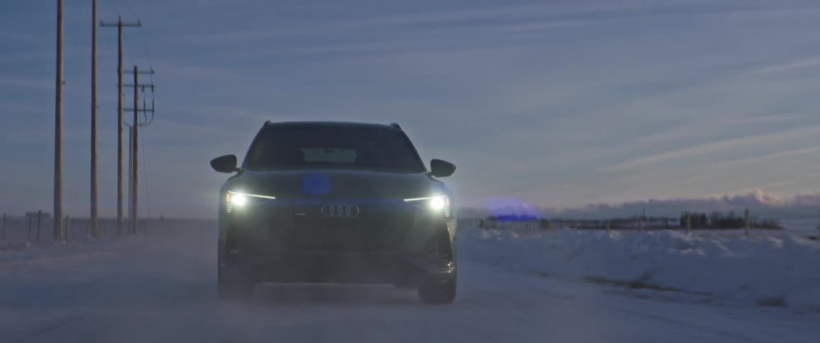 A black car travels on a snow covered road with headlights on and utility poles along the roadside