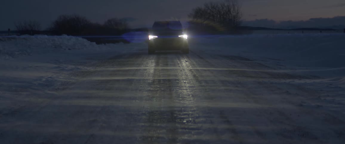 A black car with headlights on travels on a road with snow drifting across it