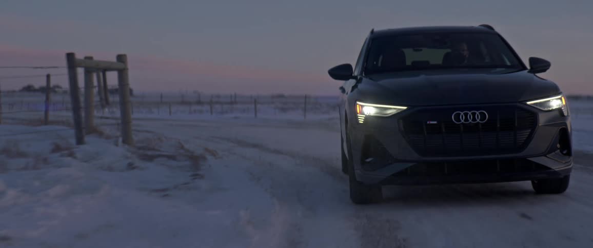 A black car travels on a snow covered road with headlights on