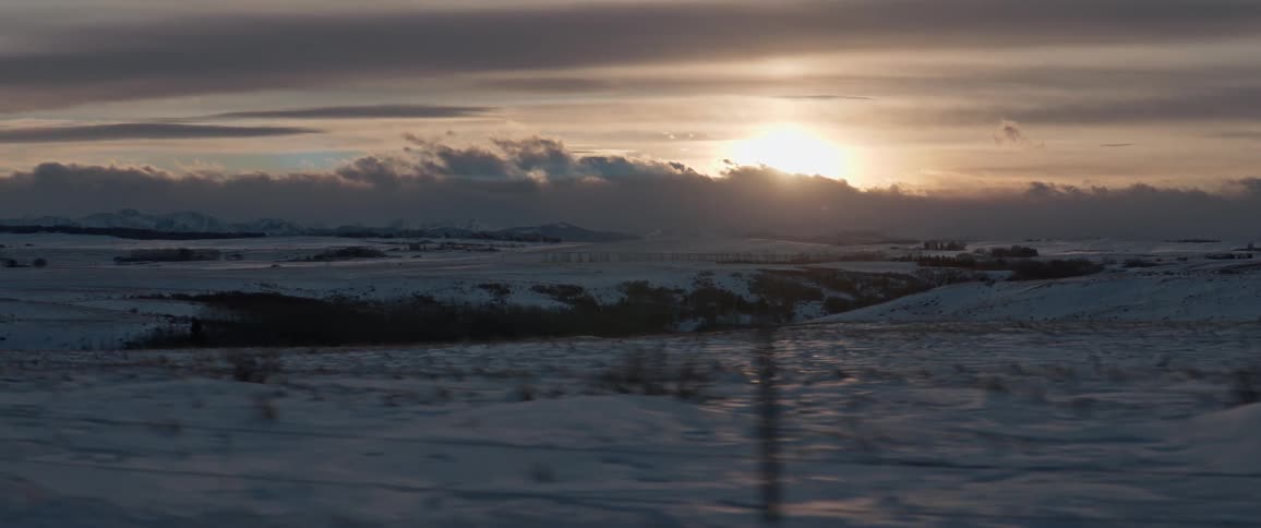 A fence with a snowy landscape with the sun the sky and clouds in the background