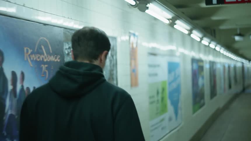 A man with a hoodie walking in a train station with posters on the wall in the background
