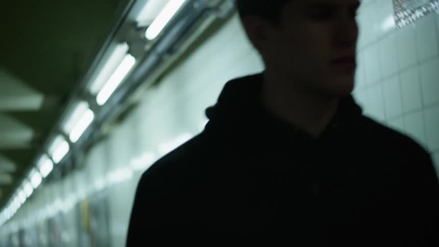 Man wearing hoodie walks down long empty corridor lit by fluorescent bulbs