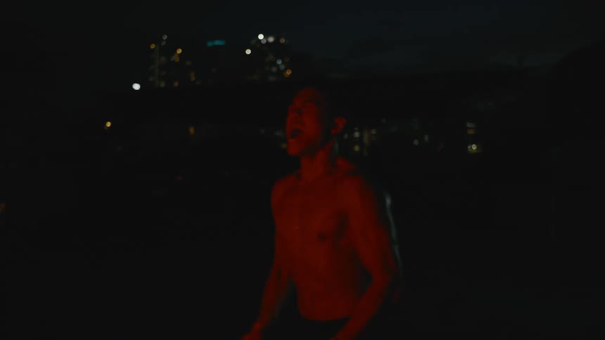 Bare chested man stands outdoors at night bathed in red light yelling with city lights in background