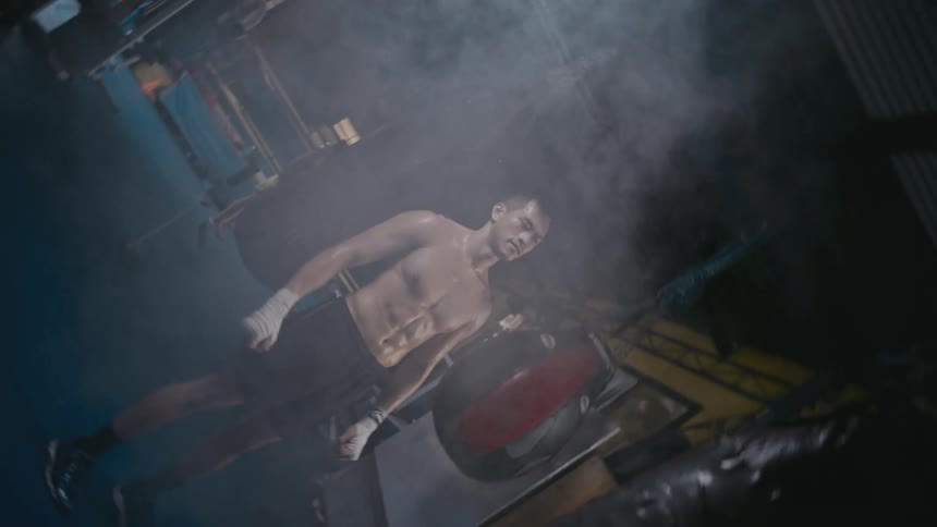 A man standing in a boxing gym with smoke with a heavy bag and a boxing ring in the background