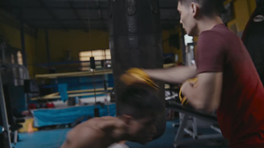 A man with boxing gloves practicing in a boxing gym with a heavy bag and a boxing ring in the background