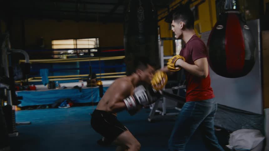 Two men in a boxing gym training with a heavy bag and a boxing ring in the background