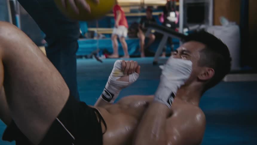 Fighter grimaces as trainer bounces medicine ball off his abdominals while people walk around near ring in background