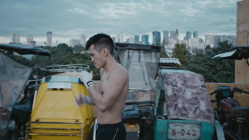 Barechested boxer walks past parked vehicles shadow boxing with city skyline on horizon under heavily clouded sky