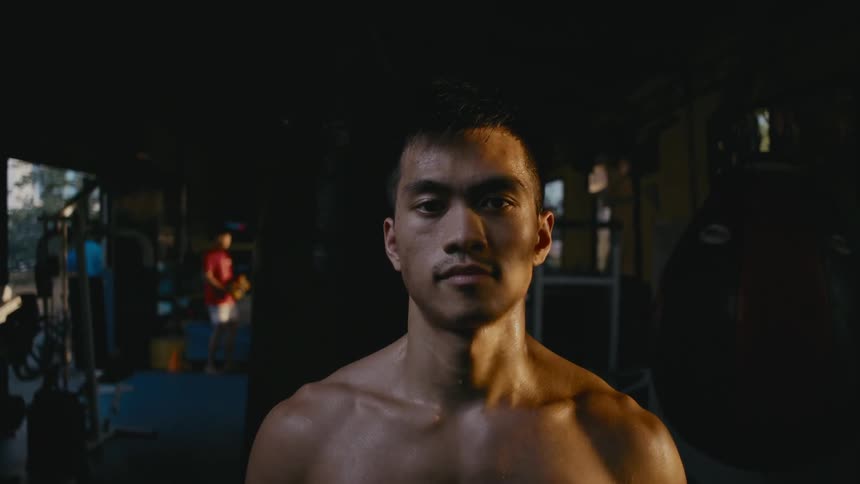 Young shirtless muscular man staring at the camera inside a dimly lit gym