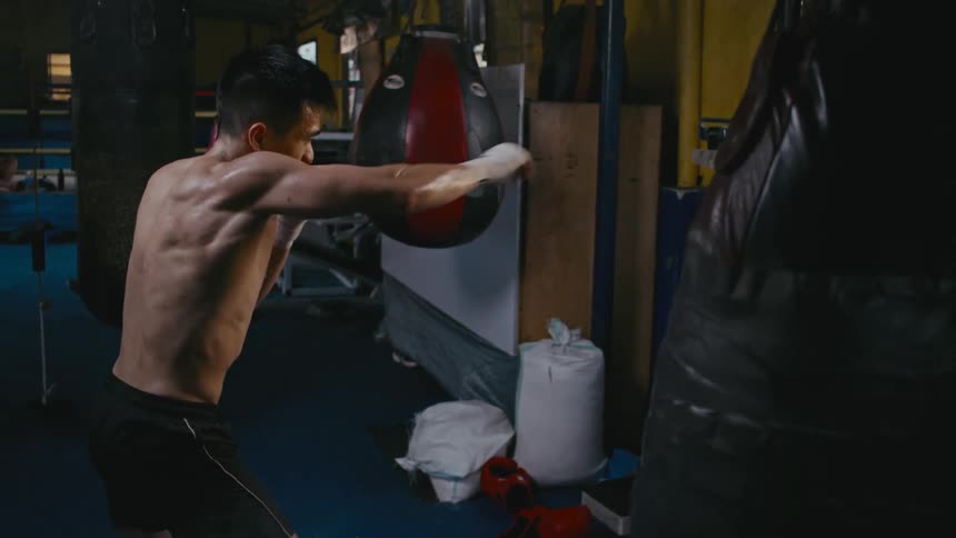 A man shadowboxing in a boxing gym with a heavy bag and a boxing ring in the background