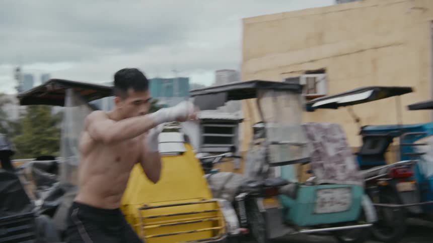A man with shorts shadowboxing on a rooftop with buildings the sky and clouds in the background