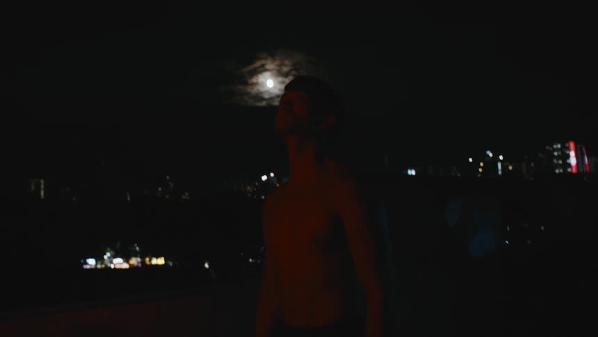 A man standing in a rooftop with buildings the moon the sky and clouds in the background