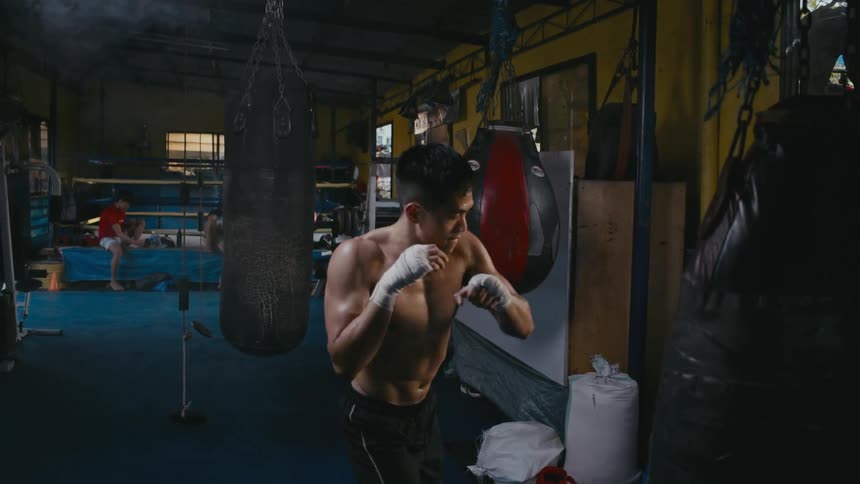 A man shadowboxing in a boxing gym with a heavy bag and a boxing ring in the background