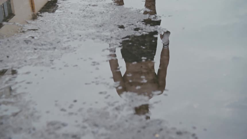 A reflection of a man with shorts in a puddle of water with the sky and clouds above
