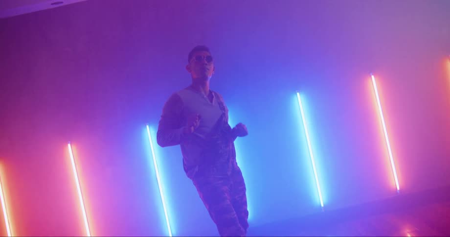 A man with sunglasses dancing in a room with a wall and lights in the background